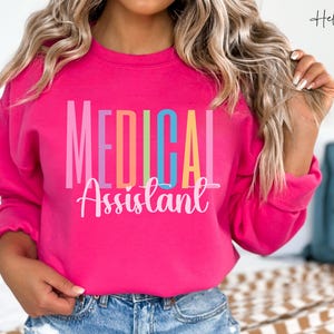 May include: A vibrant pink sweatshirt with the words "MEDICAL Assistant" in colorful block letters. The text is in pink, orange, green, and blue. The sweatshirt is a crew neck style.