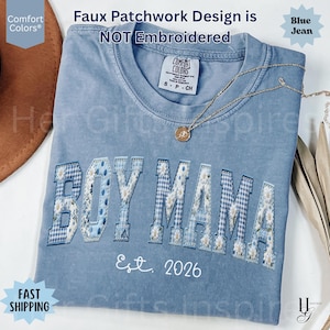 May include: A blue jean colored t-shirt with a crew neckline and a faux patchwork design featuring the text BOY MAMA and Est. 2026. The letters have a blue and white gingham and floral pattern. A gold-toned necklace rests on the shirt collar.
