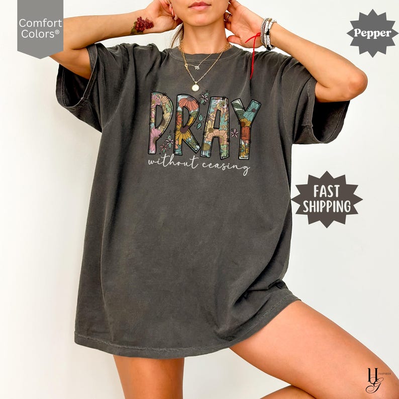 May include: A dark gray Comfort Colors t-shirt with the word "PRAY" in a floral design, and the words "without ceasing" below. The shirt has short sleeves and a relaxed fit. The word "Pepper" is in the top right corner.