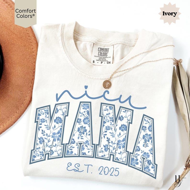 May include: Ivory-colored Comfort Colors t-shirt with the words "nicu MAMA" in blue floral print. The text "EST. 2025" is below the word "MAMA". A gold necklace with a pendant is draped on the shirt.