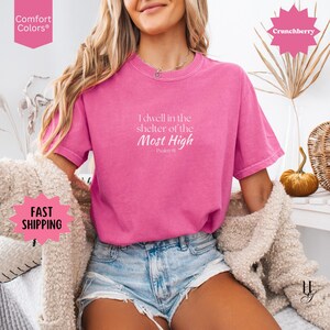 May include: A bright pink short-sleeve t-shirt featuring the white printed text I dwell in the shelter of the Most High Psalm 91 centered on the chest. The garment has a crew neckline and a relaxed fit, paired with light blue denim shorts.