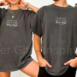 May include: A charcoal gray short-sleeve t-shirt featuring the white printed text "I dwell in the shelter of the Most High" followed by "Psalm 91" centered on the chest. The garment has a crew neckline and a relaxed, loose-fitting silhouette.