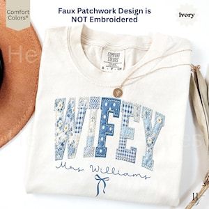 Op de afbeelding: Ivoor kleurig t-shirt met het woord "WIFEY" in een patchwork ontwerp, met blauwe bloemen- en gingham patronen. Het shirt bevat ook de naam "Mrs. Williams" en een strik afbeelding. Een gouden ketting met een hanger ligt op het shirt.