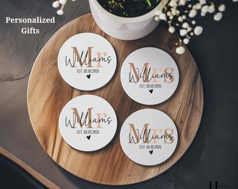Op de afbeelding: Vier witte onderzetters met een gepersonaliseerd ontwerp, met de naam "Williams" in een stijlvol lettertype, de datum "EST. 03.03.2025" en een klein hartje. De onderzetters zijn gerangschikt op een houten dienblad, met een plant in een witte pot op de achtergrond.
