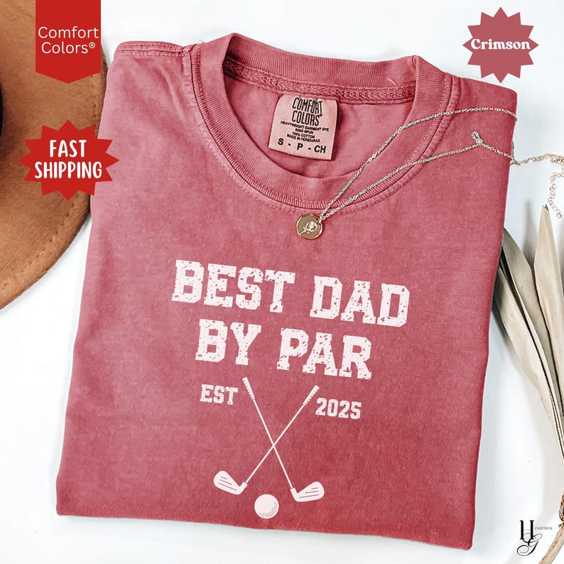 Peut inclure: T-shirt couleur pourpre avec l'inscription blanche "BEST DAD BY PAR EST 2025". Le motif comprend des clubs de golf crois&eacute;s et une balle. Un collier argent&eacute; avec un pendentif est pos&eacute; sur le t-shirt.