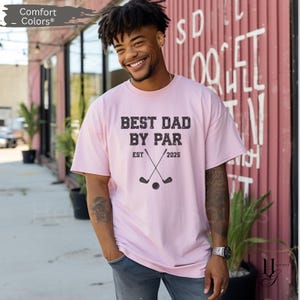 Peut inclure: T-shirt rose clair avec l'inscription "BEST DAD BY PAR EST 2025" en gris fonc&eacute;. Le motif comprend des clubs de golf crois&eacute;s et une balle de golf. Marque Comfort Colors.