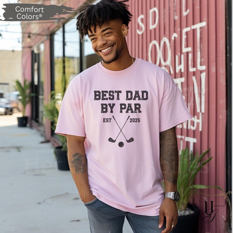 Peut inclure: T-shirt rose clair avec l'inscription "BEST DAD BY PAR EST 2025" en gris fonc&eacute;. Le motif comprend des clubs de golf crois&eacute;s et une balle de golf. Marque Comfort Colors.