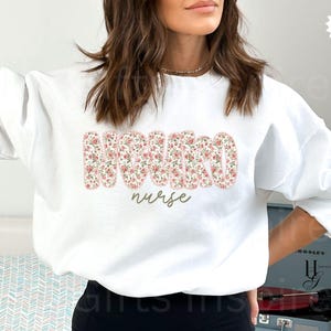 Puede incluir: Sudadera blanca con la palabra "NURSE" en un estampado floral en el pecho. La palabra "nurse" est&aacute; escrita en cursiva debajo de las letras florales. La sudadera es de cuello redondo.
