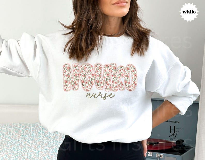 Puede incluir: Sudadera blanca con la palabra "NURSE" en un estampado floral en el pecho. La palabra "nurse" est&aacute; escrita en cursiva debajo de las letras florales. La sudadera es de cuello redondo.