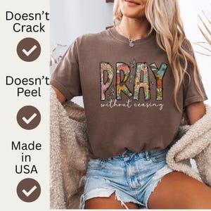 May include: A brown t-shirt with the word "PRAY" in a floral design, with the words "without ceasing" underneath. The shirt is paired with denim shorts. The image also includes text that says "Doesn't Crack", "Doesn't Peel", and "Made in USA".