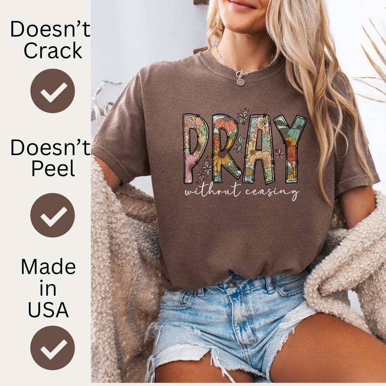 May include: A brown t-shirt with the word "PRAY" in a floral design, with the words "without ceasing" underneath. The shirt is paired with denim shorts. The image also includes text that says "Doesn't Crack", "Doesn't Peel", and "Made in USA".