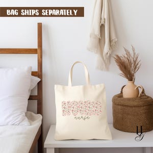 Puede incluir: Bolso tote de color crema con la palabra "NURSE" en un estampado floral. La palabra "nurse" est&aacute; escrita en cursiva debajo. El bolso tiene dos asas y est&aacute; sobre una mesa blanca. El texto "BAG SHIPS SEPARATELY" est&aacute; arriba.