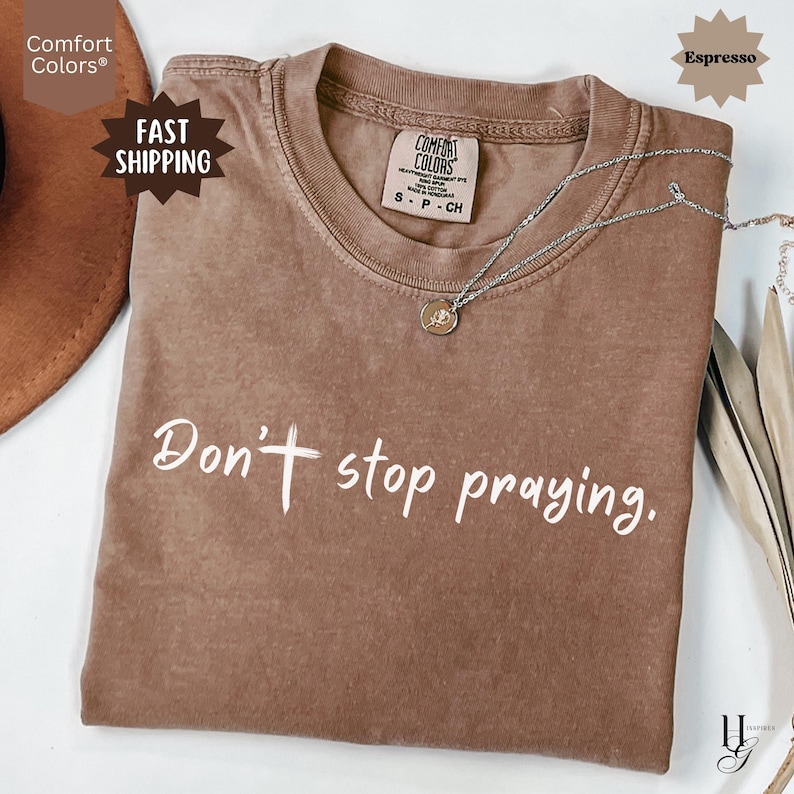 May include: Espresso-colored Comfort Colors t-shirt with the phrase "Don't stop praying" in white script. A silver necklace with a small pendant rests on the shirt. The shirt has a crew neck and short sleeves.