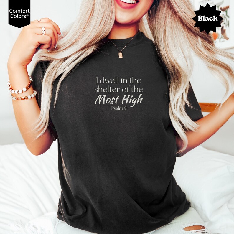 May include: A black short-sleeve t-shirt featuring white text that reads I dwell in the shelter of the Most High Psalm 91. The shirt has a crew neckline and a relaxed fit, with the Comfort Colors brand logo visible in the top left corner.