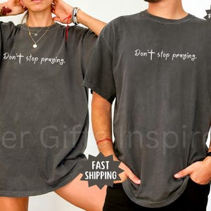 May include: Two dark gray t-shirts with the text "Don't stop praying" in white script. The shirts are a relaxed fit and have short sleeves. The image includes the words "Comfort Colors" and "Fast Shipping".