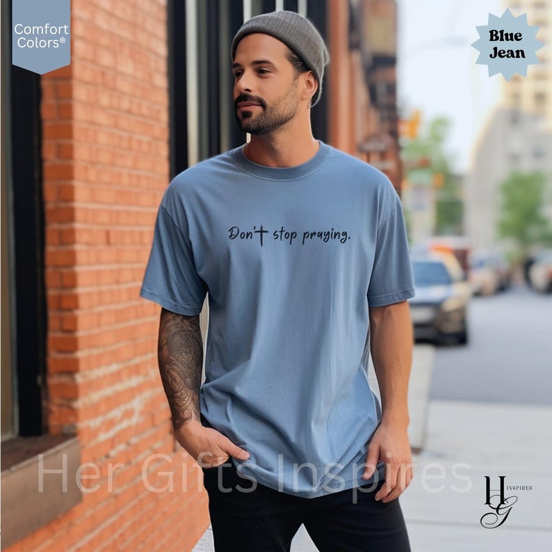 May include: Blue jean Comfort Colors t-shirt with the text "Don't stop praying." The shirt is a light blue color and has a relaxed fit. The shirt is being worn outdoors.