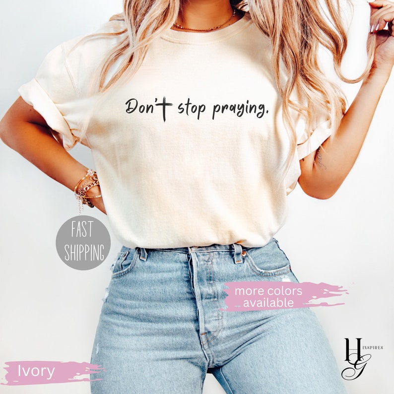 May include: Ivory-colored t-shirt with the text "Don't stop praying" in black. The shirt is paired with light blue jeans. Additional text includes "more colors available" and "Fast Shipping".