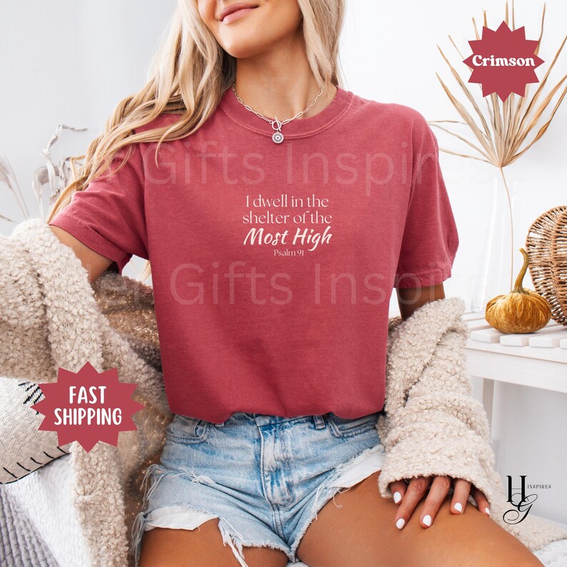 May include: A crimson short-sleeved t-shirt featuring white text that reads I dwell in the shelter of the Most High Psalm 91. The shirt has a crew neckline and is worn with light blue denim shorts and a textured beige cardigan draped over the shoulders.