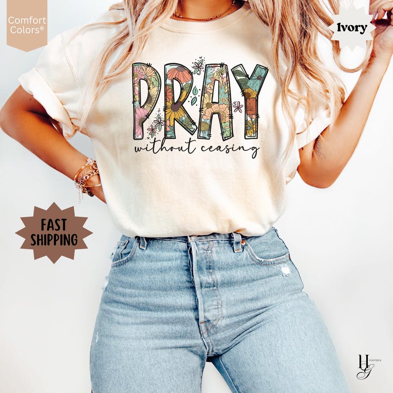 May include: Ivory-colored t-shirt with the word "PRAY" in large, floral-filled letters. Below, the phrase "without ceasing" is written in a delicate script. The shirt is paired with blue jeans. The words "Comfort Colors" and "Fast Shipping" are also visible.