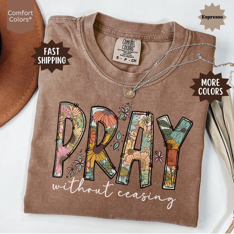 May include: Espresso-colored t-shirt with the word "PRAY" in a floral design, and the words "without ceasing" below. The shirt has a crew neck and short sleeves. A silver necklace with a pendant is on the shirt.