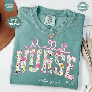 Può includere: Una maglietta verde chiaro con un design floreale che dice "Mrs. Nurse" in lettere rosa. Il nome "Abigail. RN" è scritto sotto il design floreale.