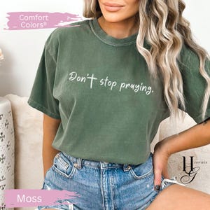 May include: Moss green Comfort Colors t-shirt with the text "Don't stop praying" in white script. The shirt has a relaxed fit and is paired with denim shorts. The shirt is a solid color and has a crew neck.