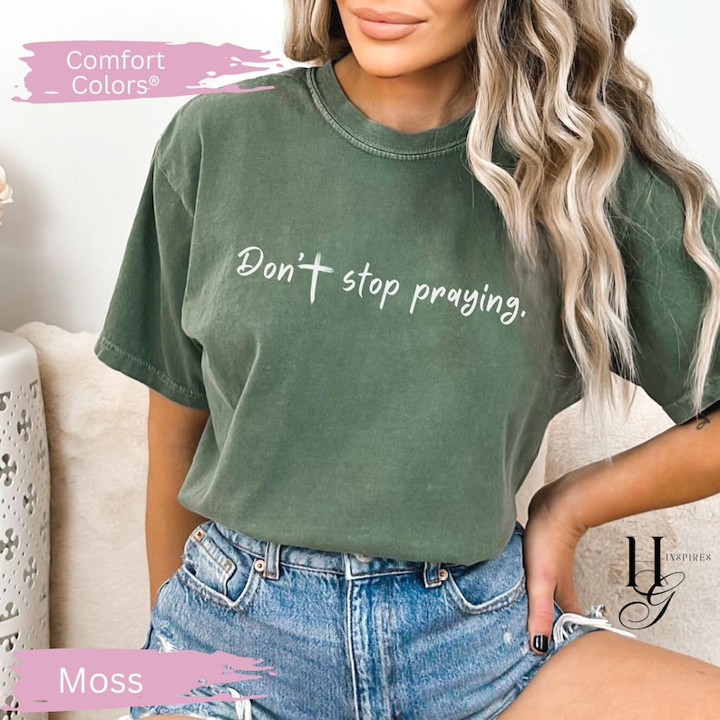 May include: Moss green Comfort Colors t-shirt with the text "Don't stop praying" in white script. The shirt has a relaxed fit and is paired with denim shorts. The shirt is a solid color and has a crew neck.