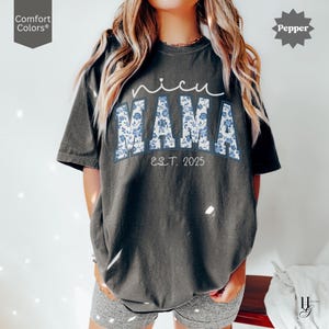 May include: Dark gray t-shirt with the text "nicu MAMA" in white script and blue floral block letters. The text "EST. 2025" is below the word "MAMA". The shirt has short sleeves and a relaxed fit.