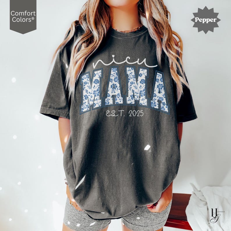 May include: Dark gray t-shirt with the text "nicu MAMA" in white script and blue floral block letters. The text "EST. 2025" is below the word "MAMA". The shirt has short sleeves and a relaxed fit.