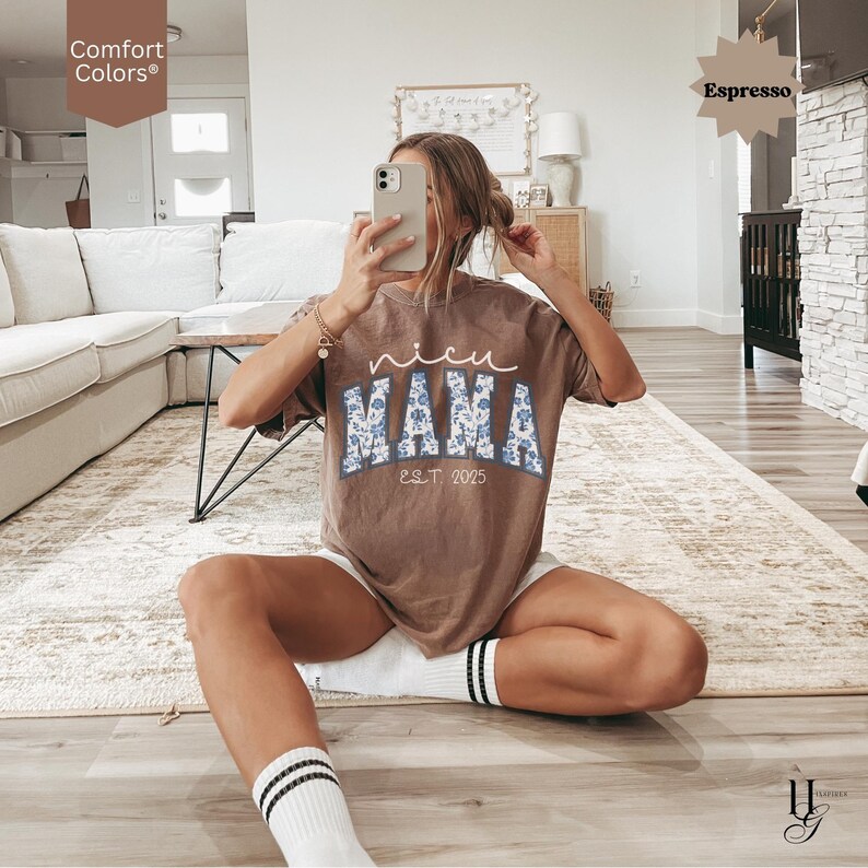 May include: A brown t-shirt with the words "nicu MAMA EST 2025" in white and blue floral print. The shirt is a Comfort Colors brand and is a relaxed fit. The model is wearing white shorts and socks with black stripes.