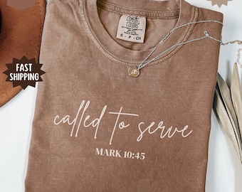 May include: An espresso-colored short-sleeve t-shirt with a crew neckline. The shirt features the white text "called to serve" in a cursive script font, followed by "MARK 10:45" in a block font. A silver-toned necklace with a small circular pendant rests on the shirt.