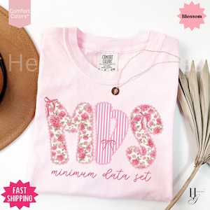 Può includere: T-shirt rosa chiaro con le lettere "MDS" in un design floreale e a righe. Il testo "minimum data set" è sotto. Una collana dorata con un ciondolo è sulla maglietta.