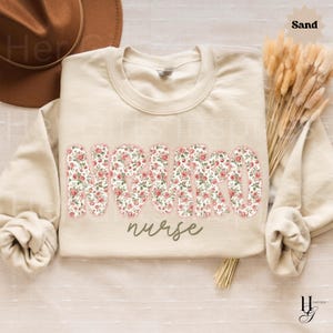 Puede incluir: Sudadera color arena con la palabra "NURSE" en letras grandes con estampado floral. La palabra "nurse" est&aacute; escrita en cursiva debajo de la palabra m&aacute;s grande. La sudadera se muestra con un sombrero marr&oacute;n y flores secas.