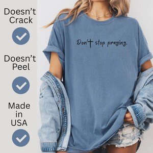 May include: A blue t-shirt with the phrase "Don't stop praying" printed in black. The shirt has rolled-up sleeves and is made in the USA. The text "Doesn't Crack" and "Doesn't Peel" are also printed on the shirt.