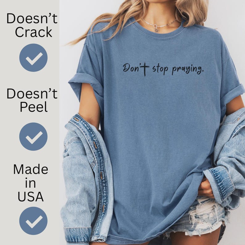 May include: A blue t-shirt with the phrase "Don't stop praying" printed in black. The shirt has rolled-up sleeves and is made in the USA. The text "Doesn't Crack" and "Doesn't Peel" are also printed on the shirt.
