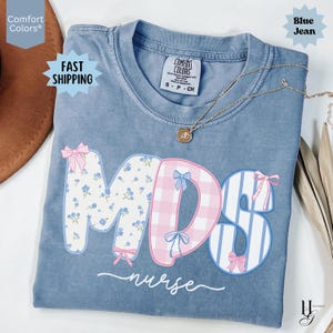 May include: Blue jean colored t-shirt with the letters "MDS" in floral, plaid, and striped patterns, accented with bows. The word "nurse" is written in cursive below the letters. A gold necklace with a pendant is draped across the shirt.