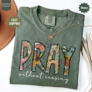 May include: A moss green t-shirt with the word "PRAY" in large, floral-filled letters. Below, the text reads "without ceasing" in a script font. A gold necklace with a pendant rests on the shirt.