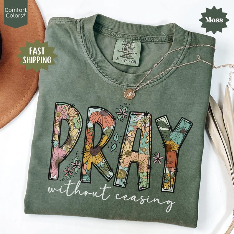 May include: A moss green t-shirt with the word "PRAY" in large, floral-filled letters. Below, the text reads "without ceasing" in a script font. A gold necklace with a pendant rests on the shirt.