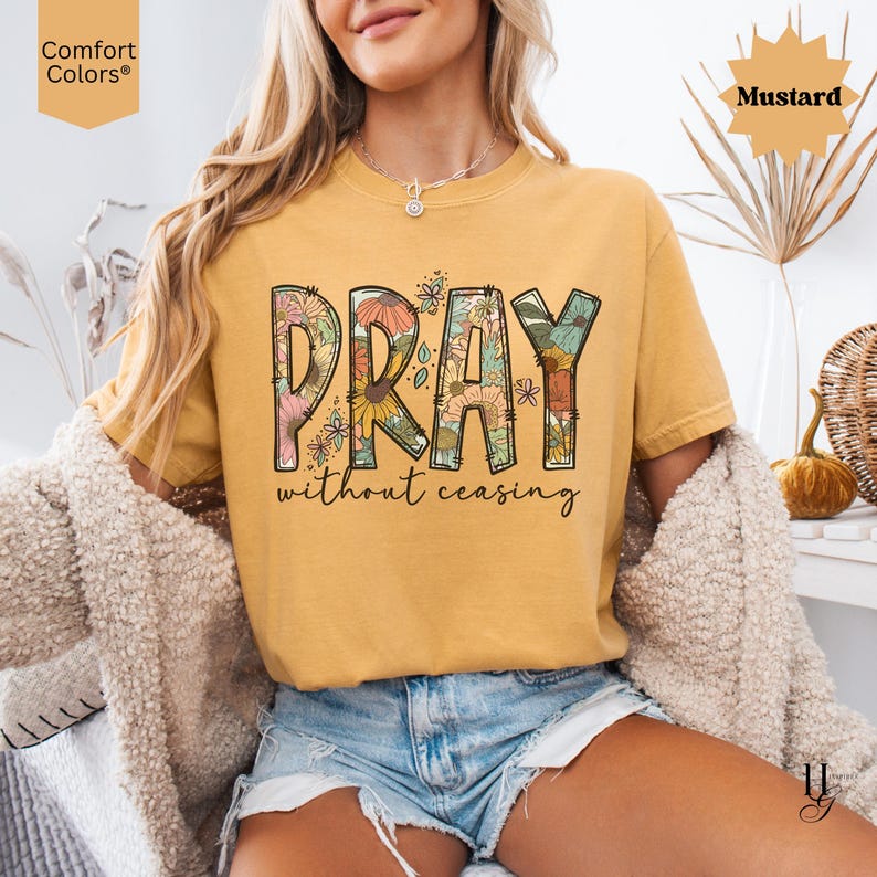 May include: Mustard-colored t-shirt with the word "PRAY" in a floral design, and the words "without ceasing" below. The shirt is a Comfort Colors brand. The model is wearing denim shorts and a cream-colored cardigan.