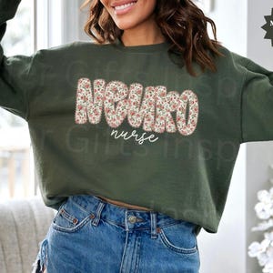 Puede incluir: Sudadera verde militar con la palabra "NURSE" en un estampado floral y la palabra "nurse" en escritura blanca. La sudadera es de manga larga y cuello redondo.