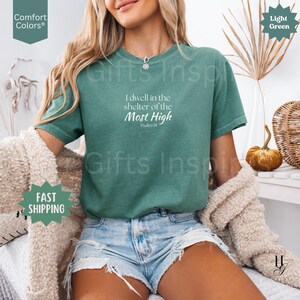 May include: A light green short-sleeve t-shirt featuring the white printed text I dwell in the shelter of the Most High Psalm 91. The shirt has a crew neckline and a relaxed fit, paired with light blue denim shorts and a beige textured cardigan.