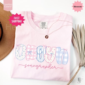Könnte beinhalten: Ein hellrosa T-Shirt mit dem Wort "OBGYN" in einer dekorativen Schriftart, mit Blumen- und Vichy-Mustern. Das Wort "Sonographer" steht darunter in Kursivschrift. Eine zarte Halskette mit Anhänger liegt auf dem Shirt.