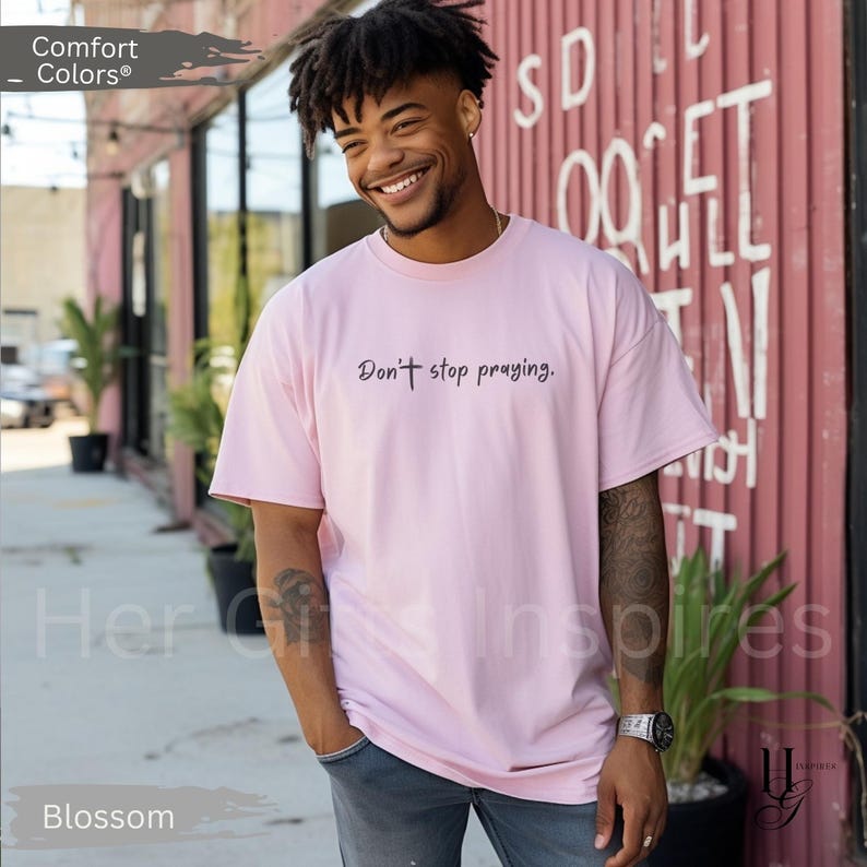 May include: A light pink t-shirt with the text "Don't stop praying." in a script font. The shirt is a Comfort Colors brand and is labeled "Blossom" at the bottom. The person is wearing blue jeans.