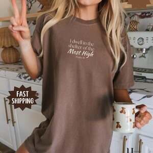 May include: An espresso-colored short-sleeve t-shirt featuring the white printed text 'I dwell in the shelter of the Most High Psalm 91' centered on the chest. The garment has a crew neckline and a relaxed fit, made from a solid-colored fabric.