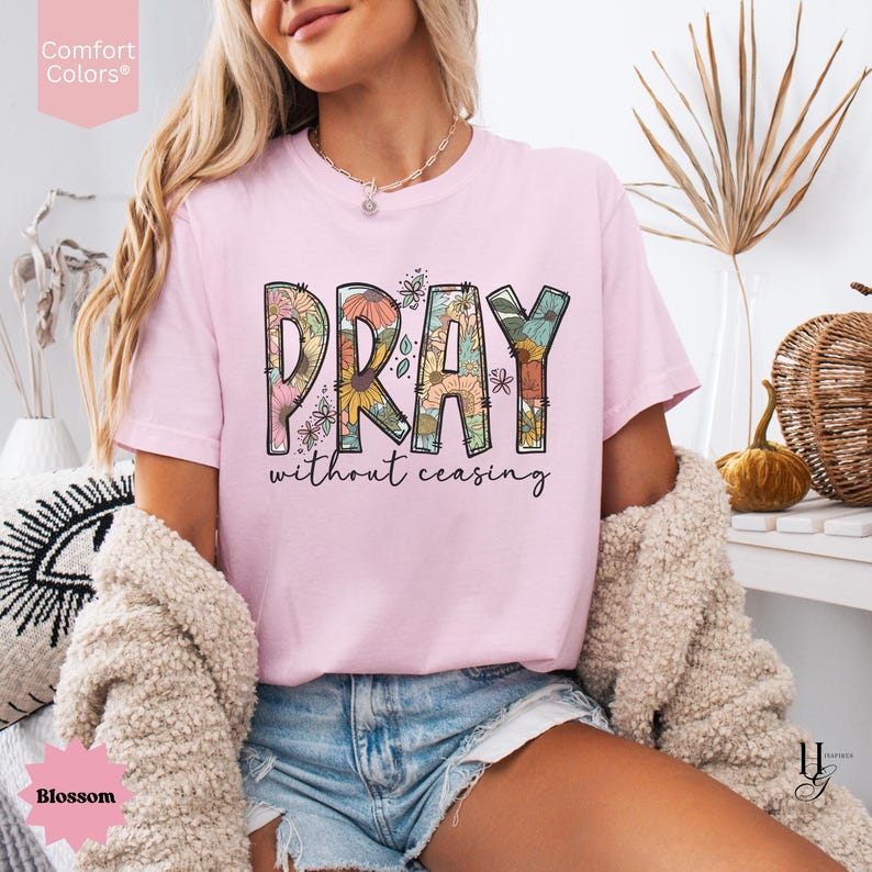May include: A light pink t-shirt with the word "PRAY" in a floral design, and the words "without ceasing" underneath. The shirt is a Comfort Colors brand and the color is Blossom.
