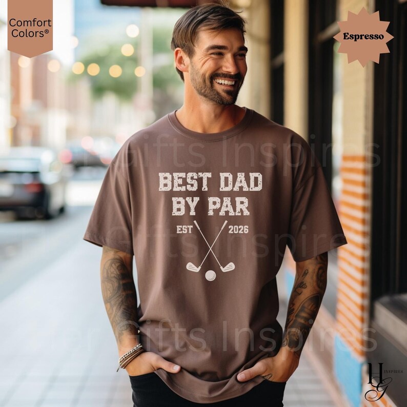 Peut inclure: T-shirt couleur expresso avec le texte "BEST DAD BY PAR" et des graphiques sur le th&egrave;me du golf. Le t-shirt comprend &eacute;galement "EST 2026". Le t-shirt est de la marque Comfort Colors.