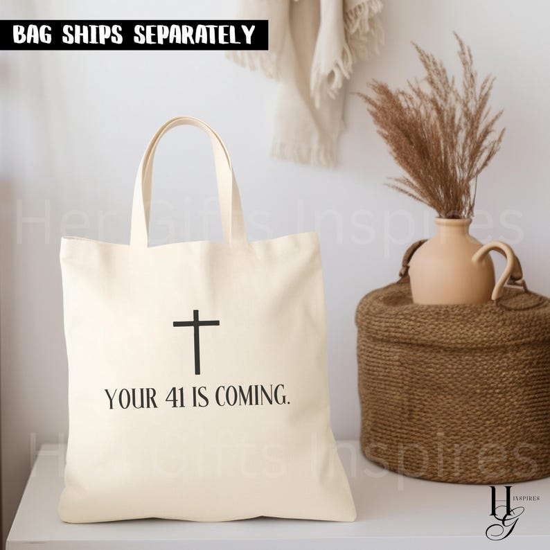 May include: A cream-colored canvas tote bag featuring a black cross graphic centered above the black text "YOUR 41 IS COMING." The bag has two shoulder straps and a simple rectangular shape. The design is printed clearly on the front of the fabric.