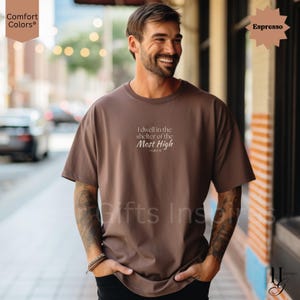 May include: An espresso-colored short-sleeve t-shirt featuring the white printed text "I dwell in the shelter of the Most High" followed by "Psalm 91" centered on the chest. The garment has a crew neckline and a relaxed, loose-fitting silhouette.