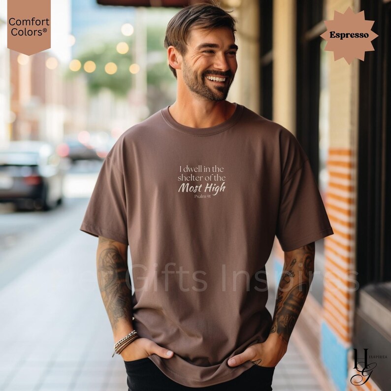 May include: An espresso-colored short-sleeve t-shirt featuring the white printed text "I dwell in the shelter of the Most High" followed by "Psalm 91" centered on the chest. The garment has a crew neckline and a relaxed, loose-fitting silhouette.