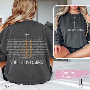 May include: A charcoal gray crewneck sweatshirt featuring a white cross and the text 'YOUR 41 IS COMING.' The back displays a detailed paragraph about the significance of the number 40 in the Bible, ending with the phrase 'YOUR 41 IS COMING.'